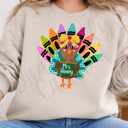 A person wearing a light beige sweatshirt with a colorful graphic of a cartoon turkey holding a chalkboard that reads 'Mrs. Honey' and surrounded by crayon feathers.