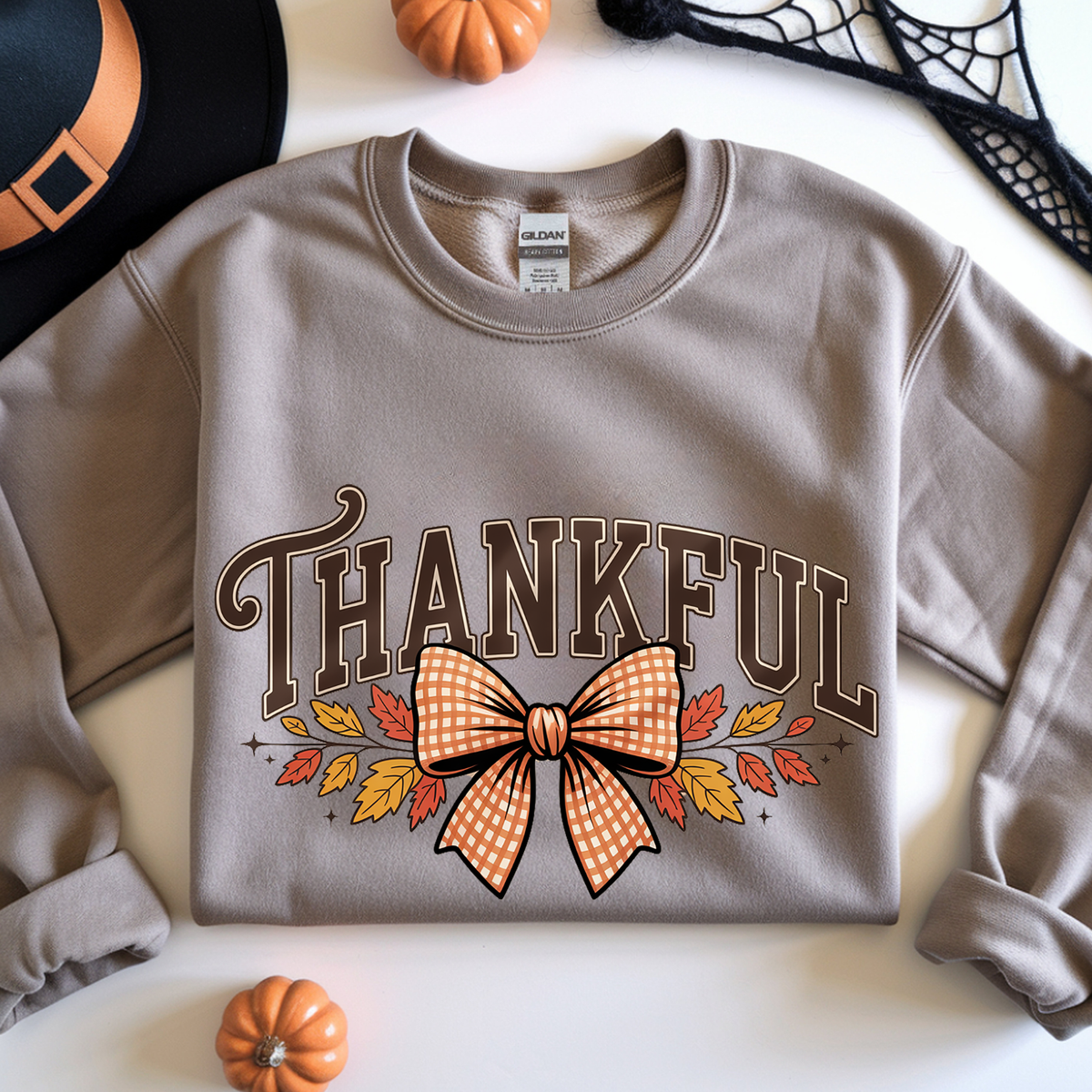 A taupe crewneck sweatshirt with a 'THANKFUL' autumn graphic featuring a gingham bow, fall leaves, and small pumpkins on a white background.