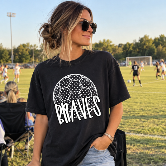 A woman wearing a black graphic t-shirt with a white soccer ball and the word 'BRAVES' stands on a grassy soccer field. The background shows a blurred soccer game with players and trees under a blue sky.