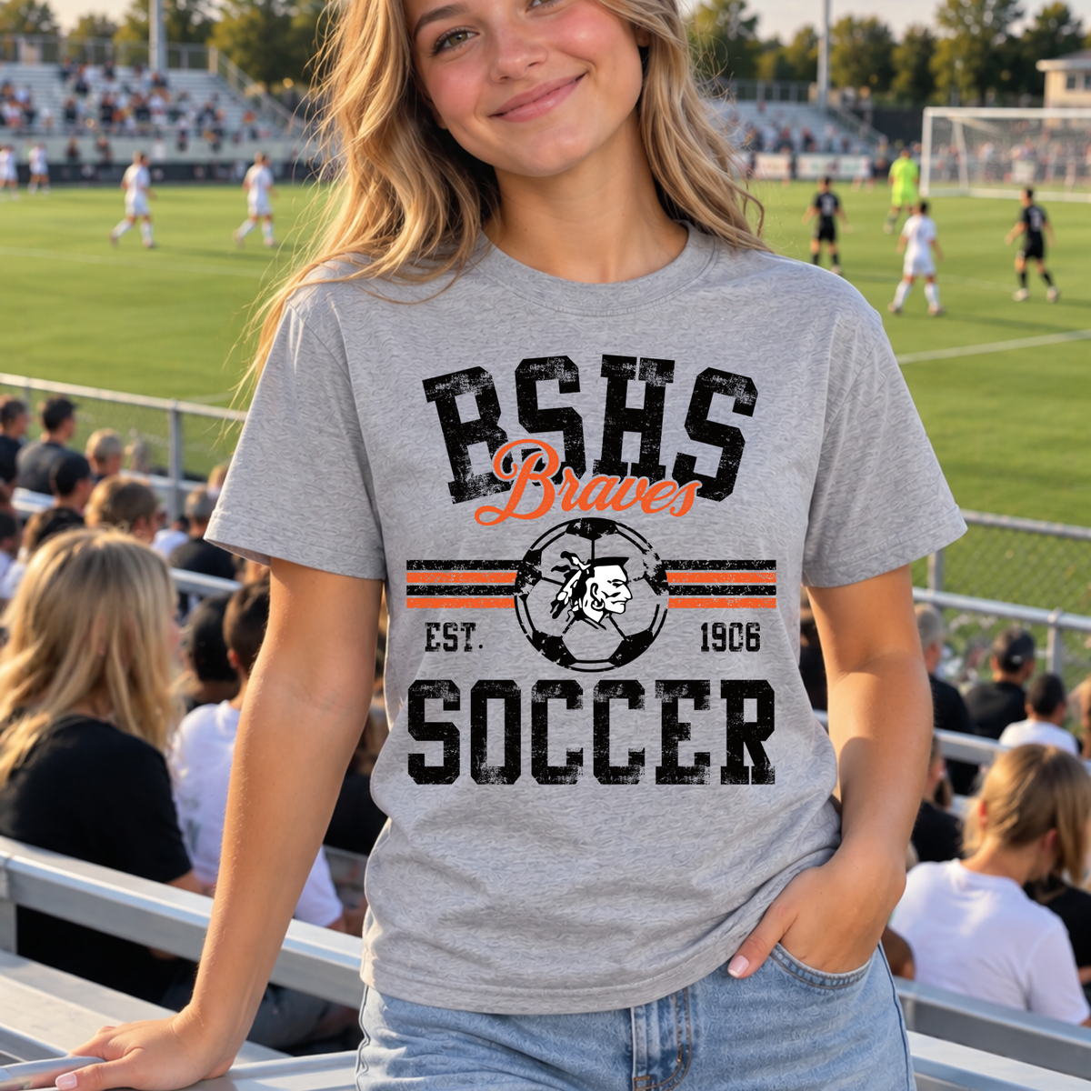 A woman wearing a heather gray graphic t-shirt with 'BSHS Braves Soccer' printed on the front, standing in a stadium with a soccer field in the background.
