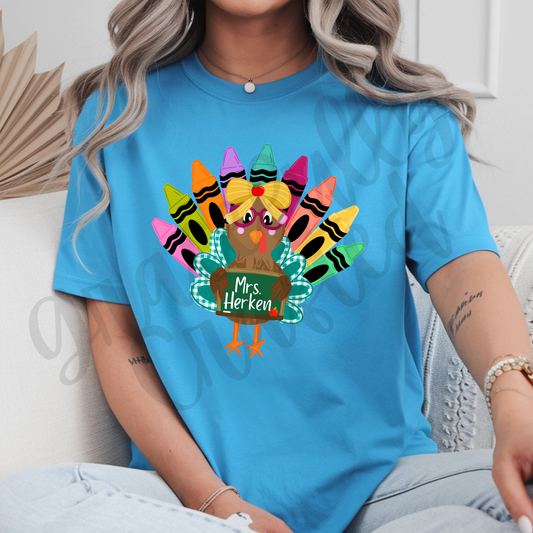 A woman wearing a bright blue t-shirt with a colorful graphic of a turkey made from crayons and a sign that reads 'Mrs. Herken'.