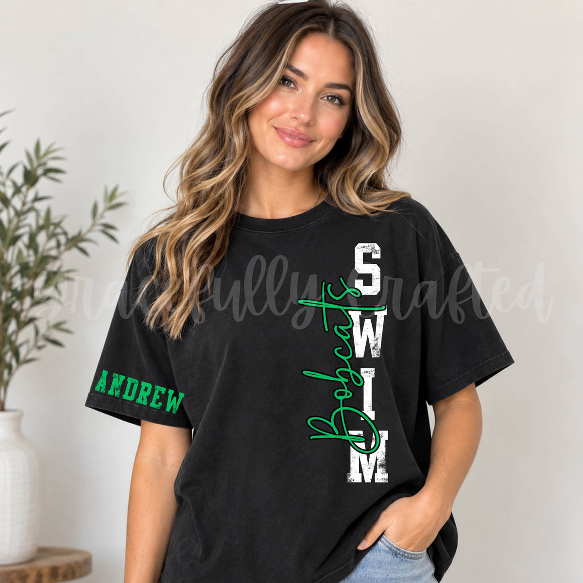 A woman wearing a black graphic t-shirt with 'BOBCATS SWIM' in white and green script on the front, and 'ANDREW' in green on the sleeve.