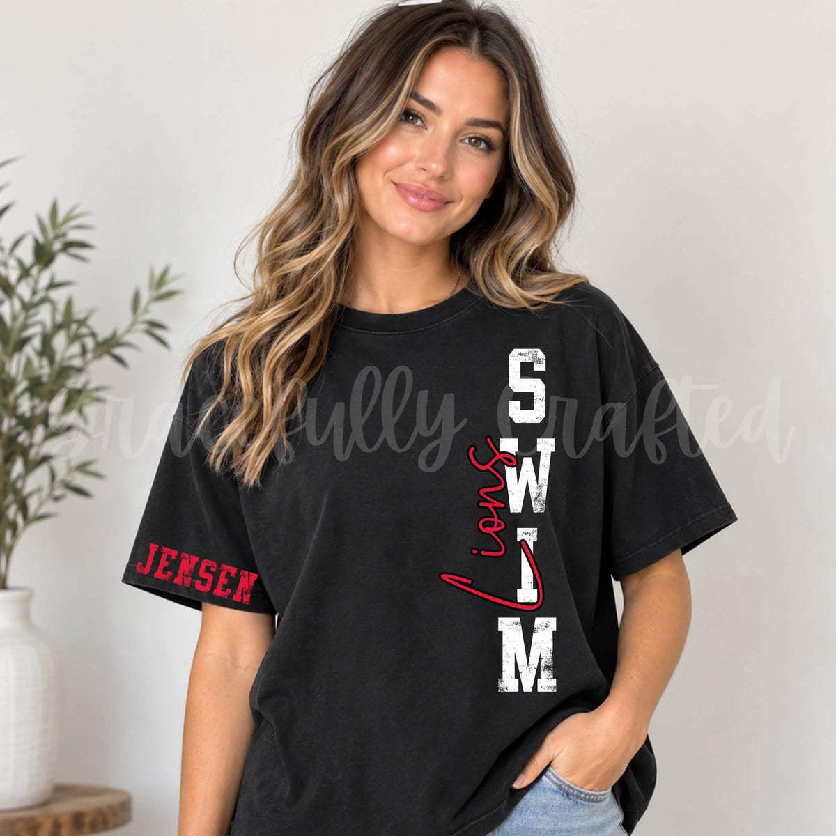 A woman wearing a black short-sleeve graphic t-shirt with 'Lions SWIM' printed vertically in white and red on the front, and 'Jensen' printed in red on the sleeve.