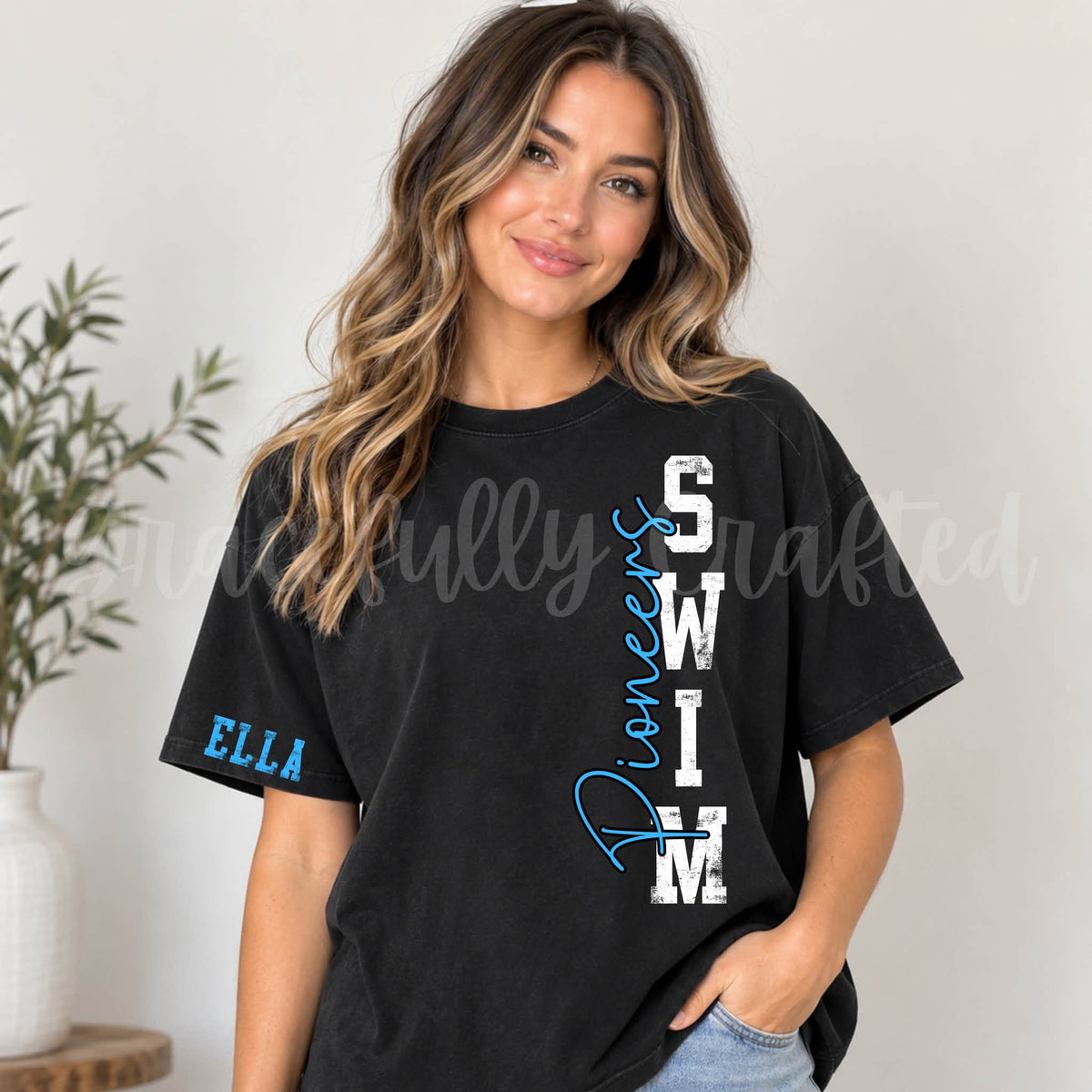 A woman wearing a black oversized t-shirt with 'Diversity Swim' printed vertically in white and blue on the front, and 'ELLA' printed in blue on the sleeve. She is posing indoors next to a potted plant.