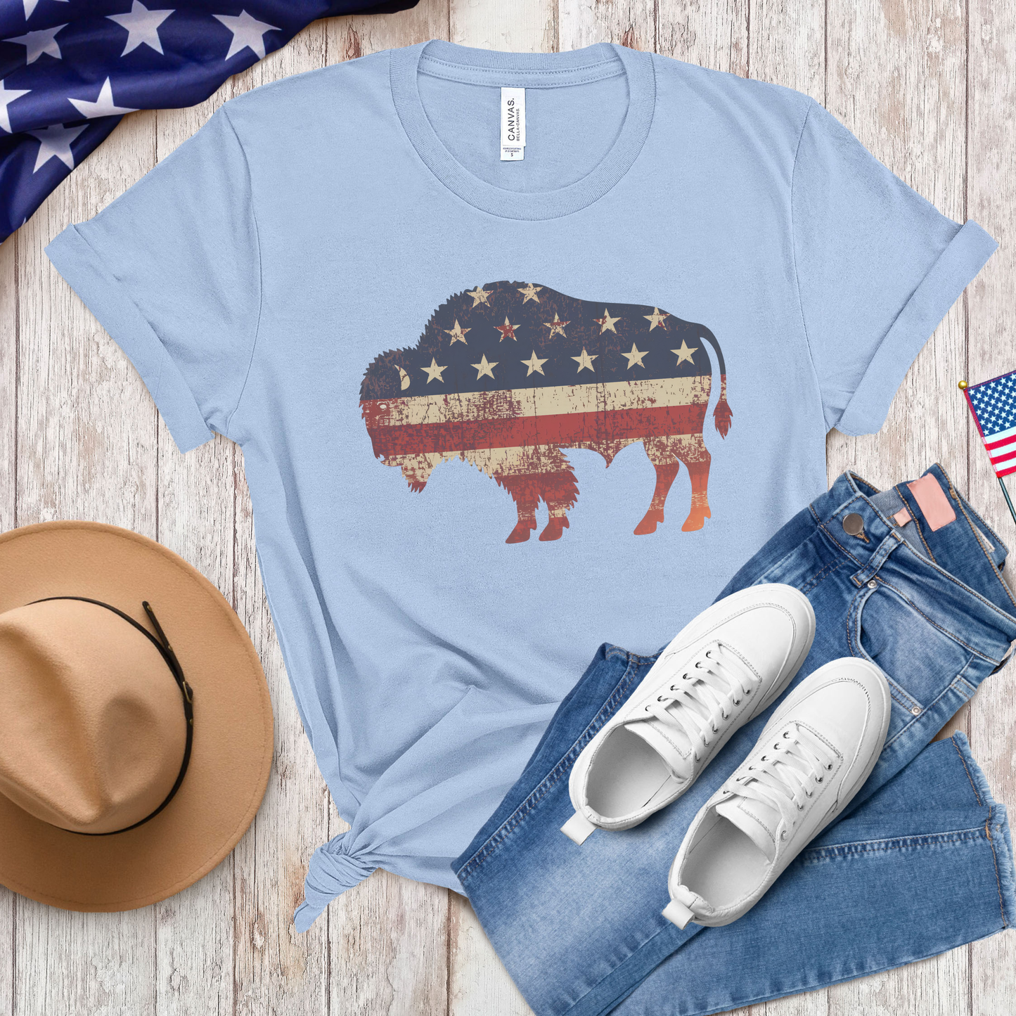 Light blue t-shirt with American flag bison design, paired with denim shorts, white sneakers, a hat, and American flags on a wooden surface.