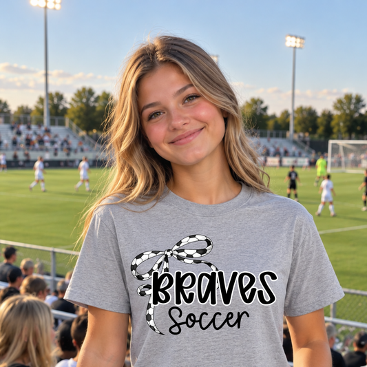 A smiling young woman stands on a soccer field at sunset wearing a heather gray t-shirt with a black and white checkered bow graphic and the words 'Braves Soccer'.