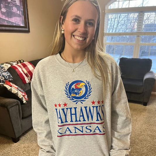 A woman wearing a heather gray crewneck sweatshirt with a Kansas Jayhawks graphic.
