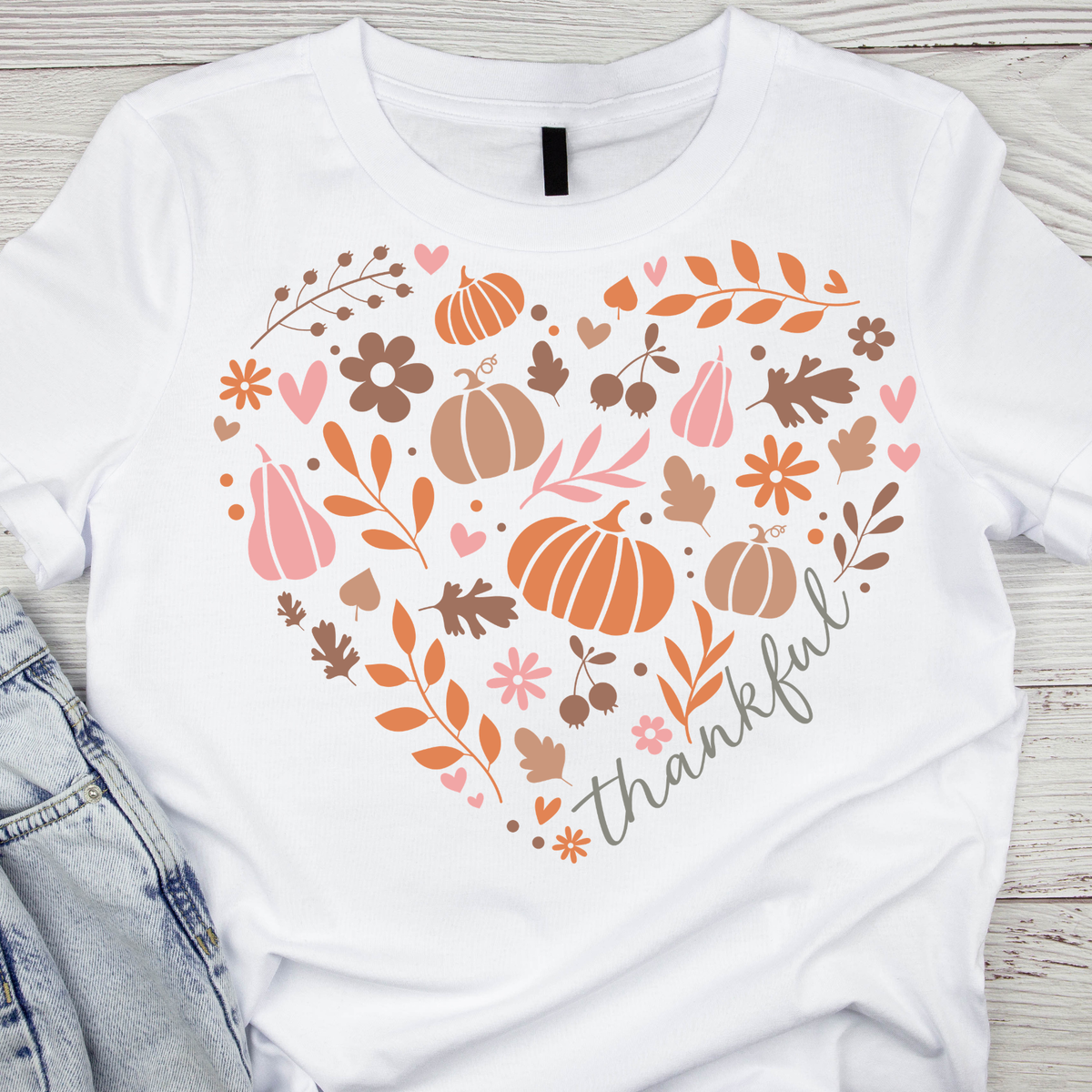 White short-sleeve T-shirt laid flat on a light wood surface, featuring a large autumn-themed heart graphic with pumpkins, leaves, flowers, hearts, and the word 'thankful.' in script.