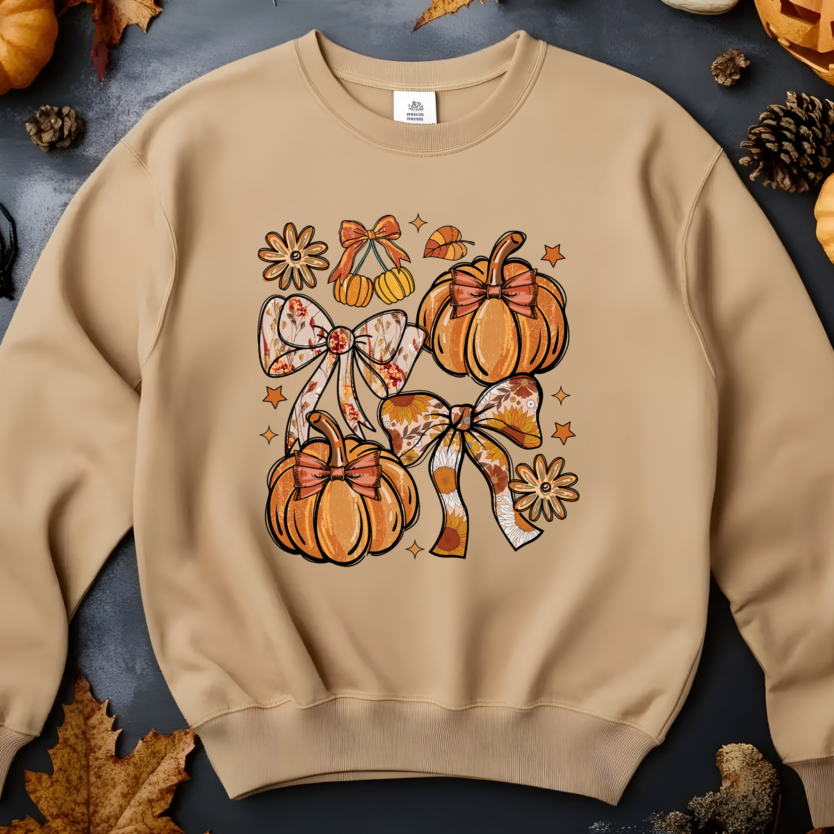 A beige crewneck sweatshirt featuring a colorful autumn graphic of pumpkins, patterned bows, and small stars, presented in a flat-lay style with seasonal props like leaves and pinecones.