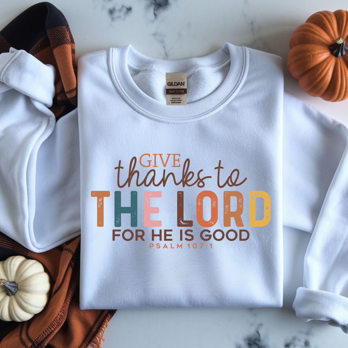White crewneck sweatshirt with a colorful Thanksgiving-themed graphic reading 'GIVE thanks to THE LORD FOR HE IS GOOD PSALM 107:1'. 