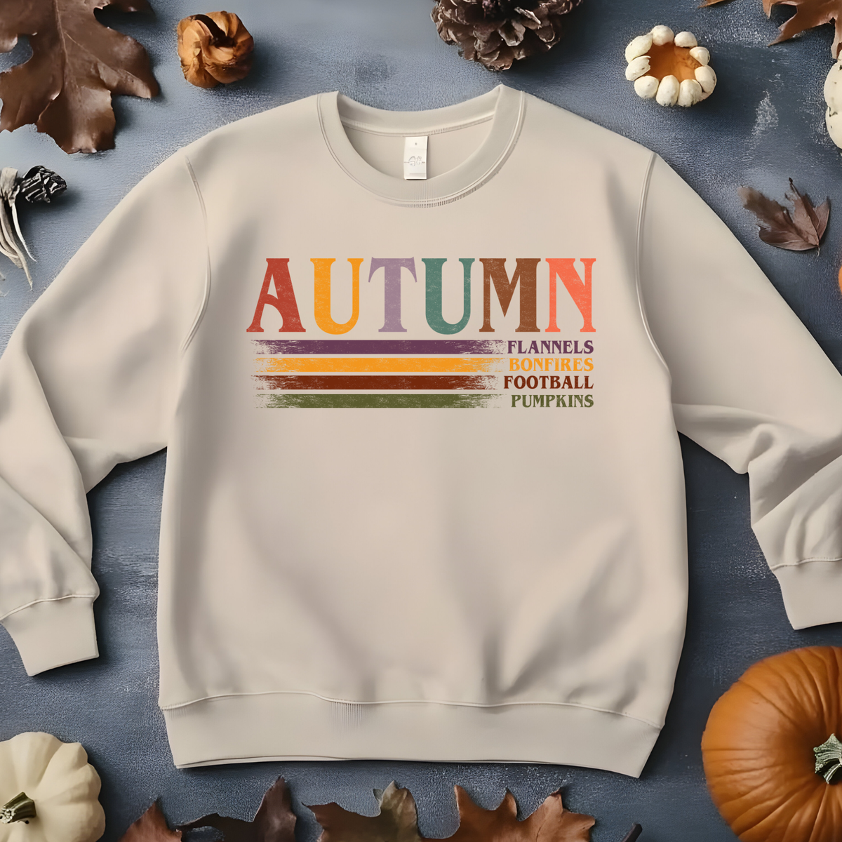 A sand-colored crewneck sweatshirt with a colorful retro graphic reading 'AUTUMN' and 'FLANNELS BONFIRES FOOTBALL PUMPKINS'