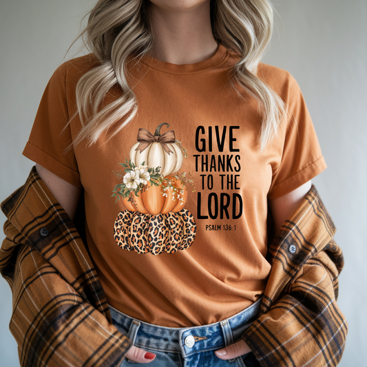 A woman wearing a rust-orange graphic t-shirt with a fall harvest design of pumpkins and the text 'GIVE THANKS TO THE LORD PSALM 106:1'. She has a brown and black plaid flannel shirt draped off her shoulders and is wearing blue jeans.