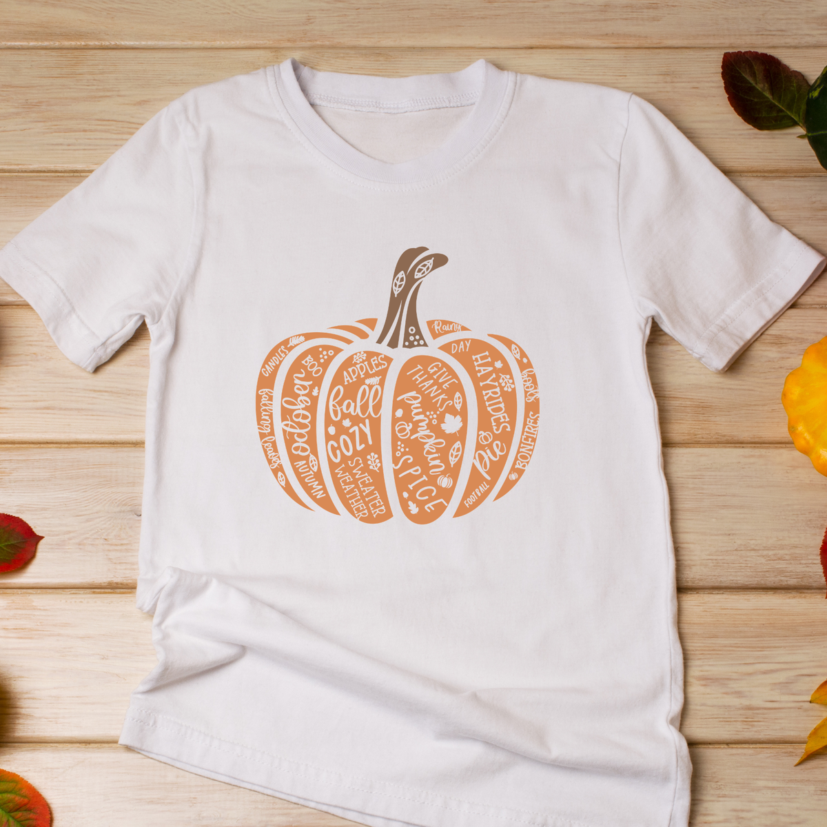 A white short-sleeve t-shirt laid flat on a light wood surface, featuring a stylized orange pumpkin graphic filled with autumn-themed words like 'Pumpkin Day', 'Fall', and 'Harvest'.