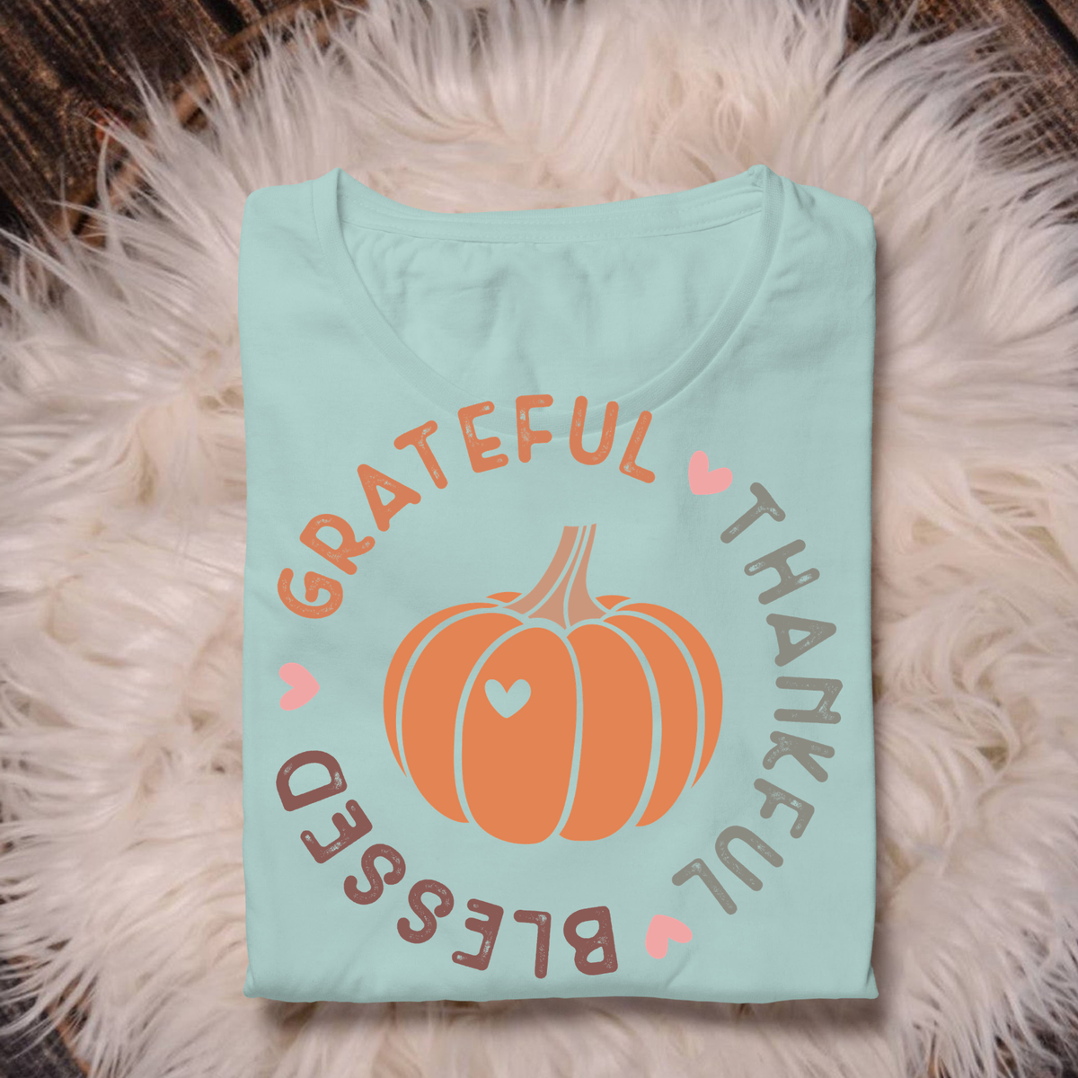 A folded light blue t-shirt with a fall-themed graphic featuring an orange pumpkin and the words 'Grateful Thankful Blessed' in peach and brown lettering