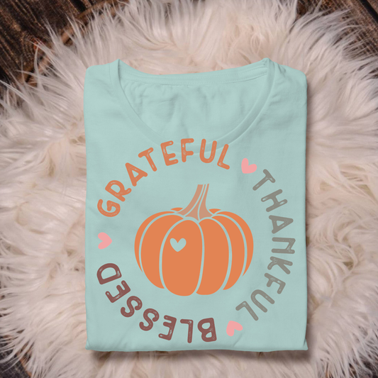 A folded light blue t-shirt with a fall-themed graphic featuring an orange pumpkin and the words 'Grateful Thankful Blessed' in peach and brown lettering