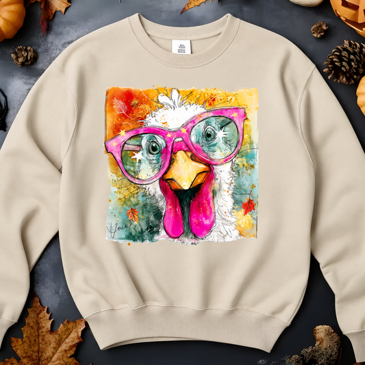 A sand crewneck sweatshirt features a large square graphic print of a white chicken wearing bright pink glasses.