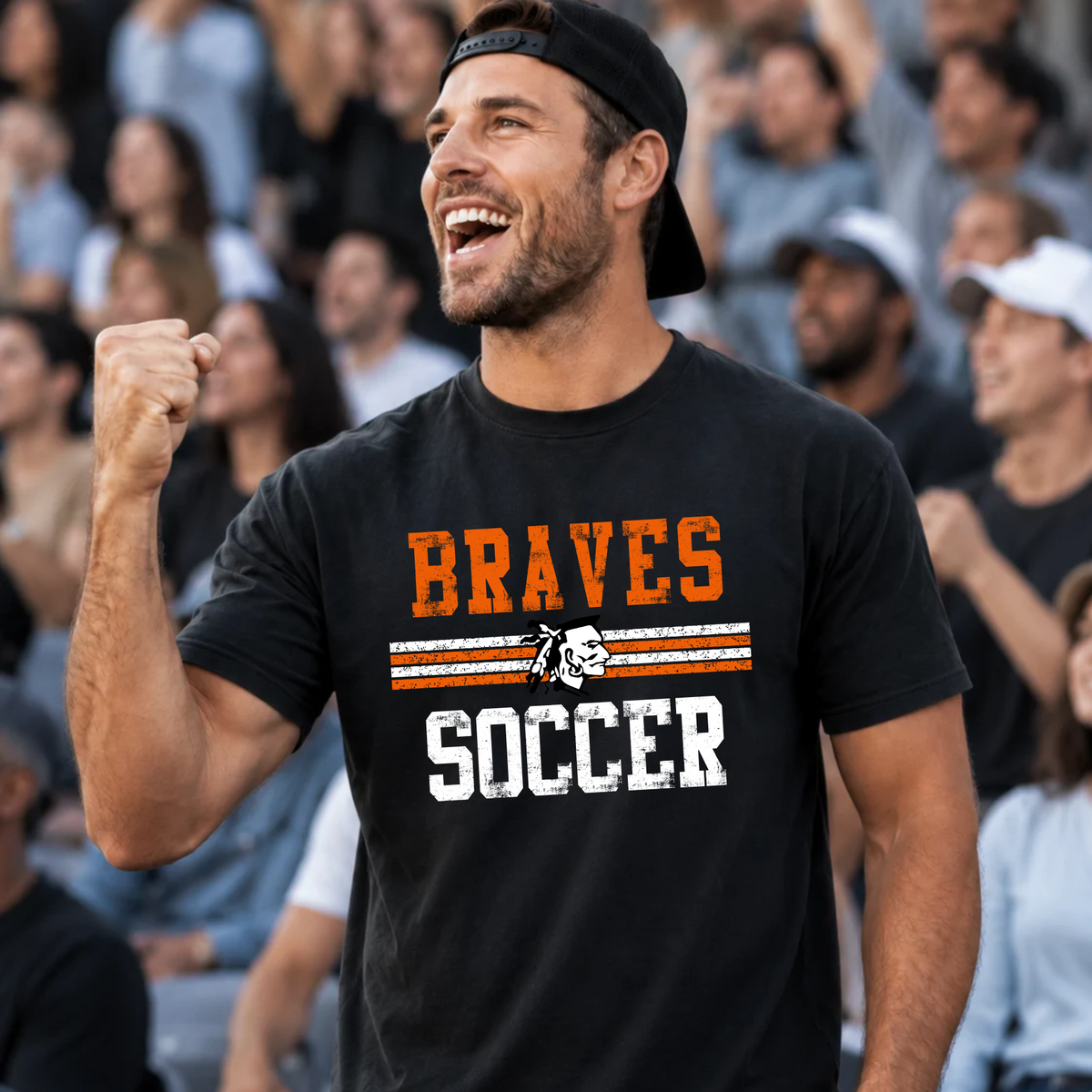 A man in a stadium wearing a black t-shirt with 'BRAVES SOCCER' printed in orange and white, along with a small soccer ball graphic.
