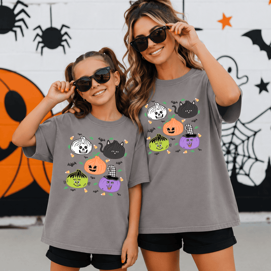 Gray Halloween t-shirt featuring five spooky pumpkin characters inspired by a skeleton, black cat, jack-o'-lantern, Frankenstein, and a purple witch wearing a checkered hat. The playful design includes scattered black bats, candy corn, and green accents.