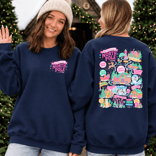 Navy blue sweatshirt featuring a small pink “Welcome to the North Pole” design on the front left chest and a colorful Christmas village map on the back. The whimsical design includes Santa’s workshop, elf cottages, reindeer, candy canes, Christmas trees, and festive North Pole destinations in bright pink, teal, green, and yellow.