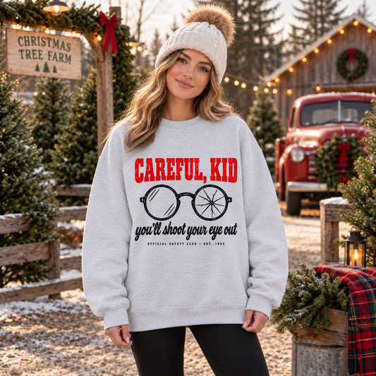 A woman wearing a light gray crewneck sweatshirt with a red and black graphic print that reads 'CAREFUL, KID you'll shoot your eye out' with glasses.